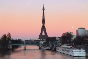 Silhouette of Eiffel tower with sunset twilight orange pink sky while full moon rising over the seine river with boats and urban modern building, trees during winter time 