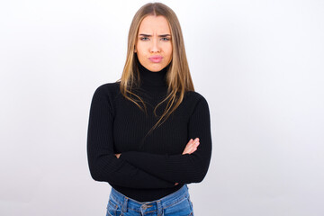 Picture of angry Young caucasian girl wearing black turtleneck over white background crossing arms. Looking at camera with disappointed expression.