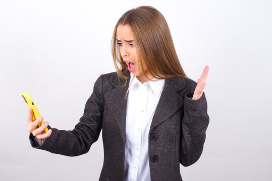 Photo of outraged annoyed Young business woman wearing jacket over white background holds cell phone, makes call, argues with colleague,  expresses negative emotions. People and anger.
