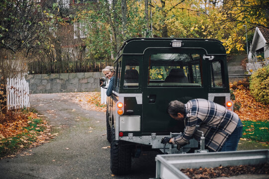 Mature man towing trailer to sports utility vehicle while female friend looking at him on driveway