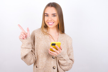 Smiling  young caucasian girl wearing knitted sweater over white background pointing finger at blank space holding phone in one hand