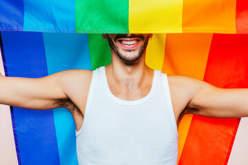 Portrait of cheerful smiling homosexual man with a gay pride flag covers part of his face at studio. Lgbtq flag, rainbow flag, celebrating parade.