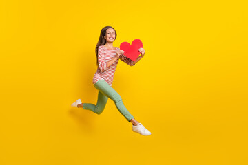 Fototapeta na wymiar Photo of cute sweet mature lady dressed striped shirt jumping walking holding big red heart smiling isolated yellow color background