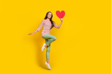 Fototapeta na wymiar Photo of pretty adorable young woman wear striped shirt smiling jumping rising large red heart empty space isolated yellow color background