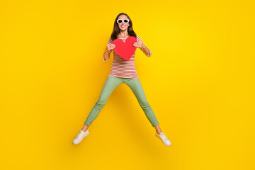 Fototapeta na wymiar Photo of sweet funny mature lady dressed striped shirt dark eyeglasses jumping high holding red heart smiling isolated yellow color background
