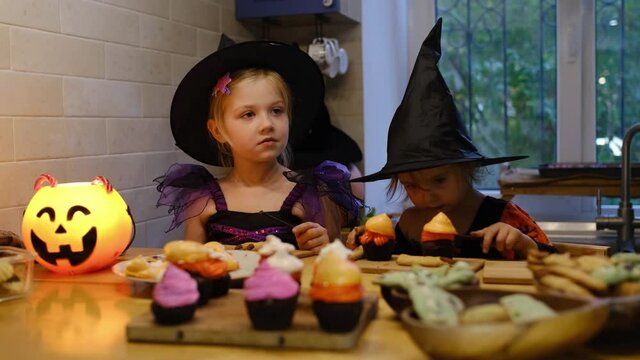 Two girls at home in the kitchen in costumes for Halloween sit with Jack-o-Lantern, laugh and taste sweets and cookies. Happy kids are getting ready for Halloween. Preparing for Halloween.
