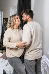 Fototapeta premium Happy family concept. Husband hug belly pregnant wife standing indoor living room near sofa