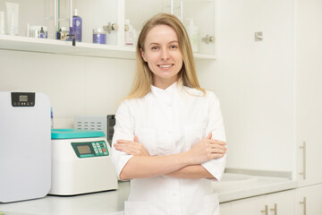 Portrait of a female doctor standing in her office at the clinic. The dermatologist beautician stands with crossed arms and looks into the camera with a smile on the background of a medical office.