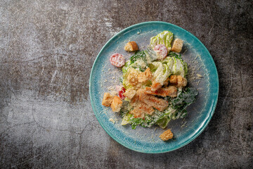 Serving a dish from the restaurant menu. Fresh Caesar salad of chicken with croutons, tomatoes and herbs on a blue plate against a gray stone table, a delicious appetizer