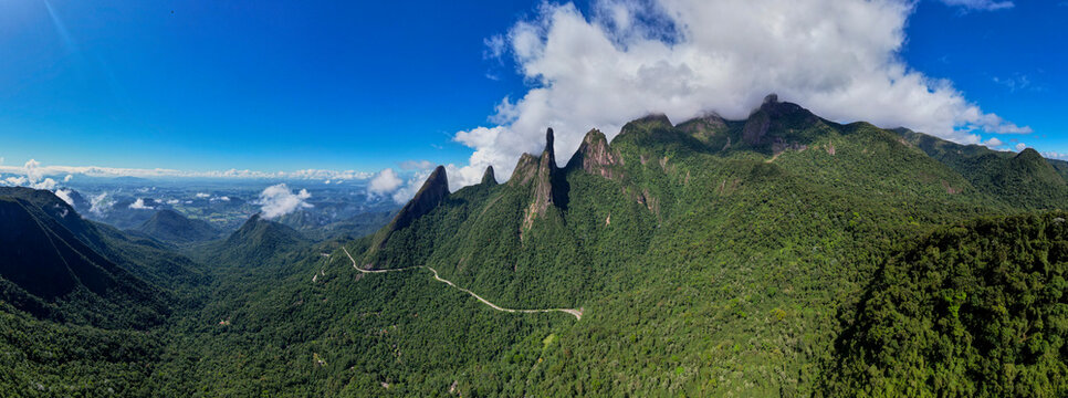 Wide aerial panorama landscape of road meandering through postcard trail Brazilian mountain range Serra dos Orgaos in Teresopolis, Rio de Janeiro, with Finger of God peak against blue sky with clouds