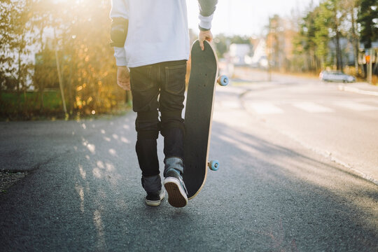 Low section of boy with skateboard walking on road