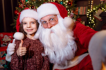 Fototapeta na wymiar Santa Claus makes selfie with charming little girl who showing thumb up. Christmas background