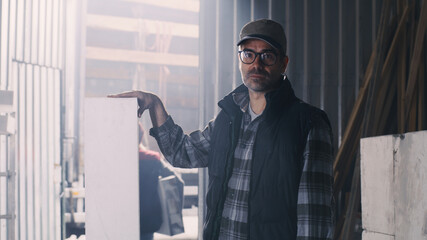 Mature male artisan with polymer block looking at camera against hangar entrance while other employees unloading truck
