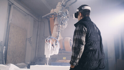 From below male worker putting on futuristic VR goggles and examining virtual plans while standing near robotic arm milling machine in illuminated workshop