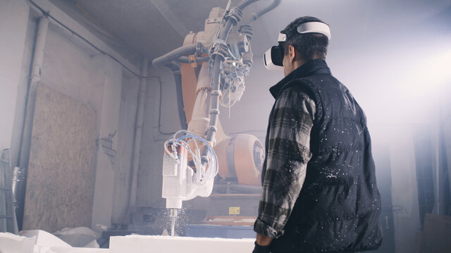 From below male worker putting on futuristic VR goggles and examining virtual plans while standing near robotic arm milling machine in illuminated workshop