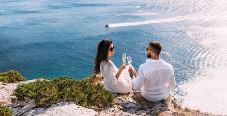 A couple in love drinking champagne on the seashore, panorama. A happy couple on a honeymoon by the sea, panorama. A beautiful couple with champagne on the coast. Honeymoon. Wedding travel. Copy space - obrazy, fototapety, plakaty