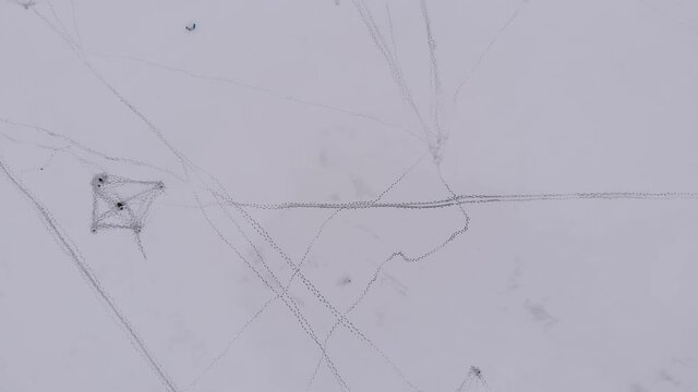Tracks of footprints of people on frozen lake covered with snow. Two fishermen fish in ice-hole on cloudy winter frosty day. Aerial view of the white surface of reservoir, tent is on the ice.