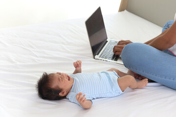 Adorable little sweet newborn baby girl crying on white bed near her mom using notebook laptop computer, cute infant with her busy mom who have to work at home.