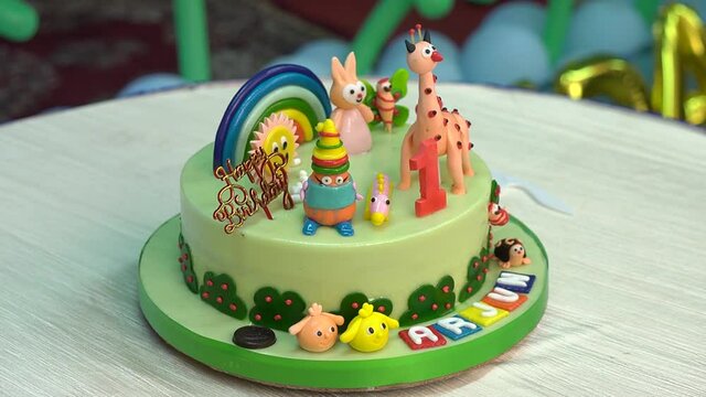 Close-up shot of a cake flavored green on top with animals, rainbow and the name written for kids birthday celebration kept on the table