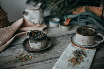 On a gray wooden table, dark ceramic mugs on saucers with herbal tea. Nearby tea towels, kettle, cinnamon and spruce branches