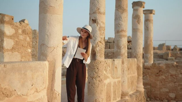 Competent female archeologist standing among ruins of ancient temple. Woman holding brush for digging and map in hands. Girl In an ancient temple with columns looking at records of a map.