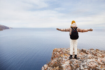 Fototapeta na wymiar A young girl traveler in warm clothes with a backpack is standing at the cliff, admiring nature. Enjoys the view of the sea or lake in late autumn. Traveling alone, healthy lifestyle, freedom. 
