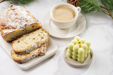 White porcelain cup with coffee cappuccino on a white saucer plate, christmas pastry stollen and a pastel green bubble candle on a marble table