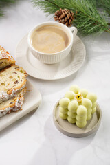 White porcelain cup with coffee cappuccino on a white saucer plate, christmas pastry stollen and a pastel green bubble candle on a marble table