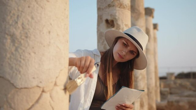 Archeologist standing among ruins of ancient temple. Woman holding brush for digging and map in hands. Girl In an ancient temple with columns looking at records of a map. Archaeological Digging Site.