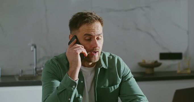 A man is talking on mobile phone, sitting at kitchen table in front of laptop