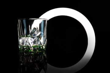 A vessel, a glass half filled with a clear liquid with ice cubes and illuminated  against a dark background with a graphic arc pattern.