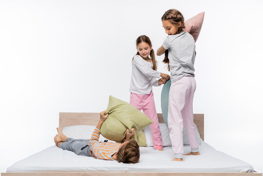 happy girls in pajamas having pillow fight while standing near boy on bed isolated on white.