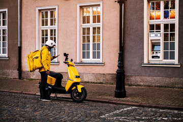 Naklejka na ściany i meble delivery man in a bright uniform on a moped picks up an order from a restaurant for quick delivery to a client. delivery man looks at the navigator in his phone