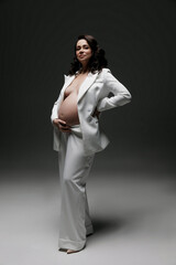 Caucasian young beautiful pregnant woman with dark hair in a white fashionable elegant pantsuit posing on a white background. isolates the banner space for text. Fashion for pregnant women. A business