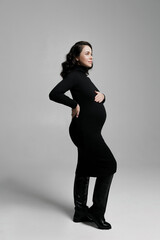 Caucasian young beautiful pregnant woman with dark hair in a tight black dress posing on a white background. elegant in a studio dress isolates the banner space for text. modern mom