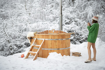 Woman taking photo of a wooden bath, during winter vacations at snowy mountains. Scandinavian lifestyle and and hot water treatments concept. Girl wearing sweater and hat