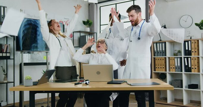 Attractive cheerful satisfied successful skilled multiethnic doctors recieving good news on laptop screen,they scattering reports over their heads,clapping hands and giving high five each other