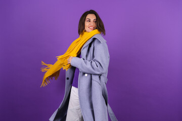 Young woman in a gray coat and a yellow scarf on a purple background posing cheerfully, cozy and warm, autumn winter style