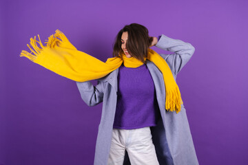 Young woman in a gray coat and a yellow scarf on a purple background posing cheerfully, cozy and warm, autumn winter style
