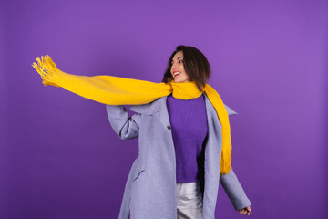 Young woman in a gray coat and a yellow scarf on a purple background posing cheerfully, cozy and warm, autumn winter style