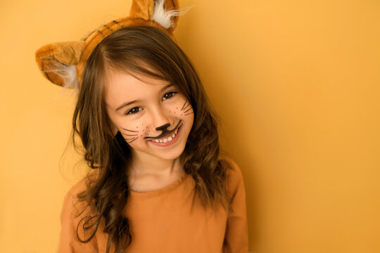 A long-haired curly-haired brunette girl with a head hoop with ears, dressed up as a tiger, smiling sweetly at the camera. Painted face of a child with a mustache of an animal for a New Year's party
