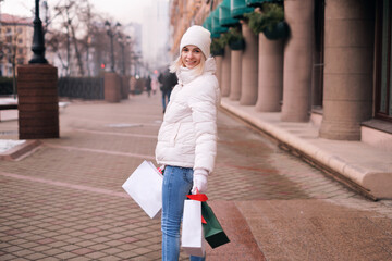 Woman in a white hat and jacket with a lot of shopping bags in hands on city streets. Columns of the store building. 