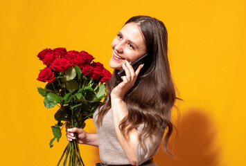 Brunette holds bouquet of red roses in one hand, and phone in other hand and smiles. Yellow background with place for text. Advertising banner for flowers, delivery. Selective focus.