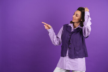 Young woman in a shirt and a vest on a purple background smiles cheerfully in high spirits, points...