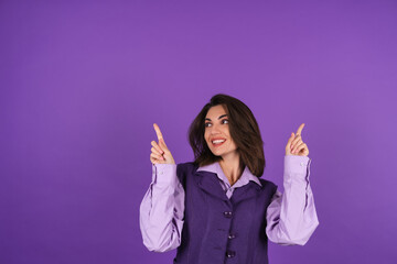 Young woman in a shirt and a vest on a purple background smiles cheerfully in high spirits, points...