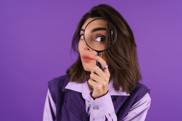 Young woman in shirt and vest on purple background having fun with magnifying glass in hand