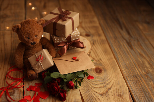 A teddy bear holds a letter and red roses on a wooden background, there are many gift boxes nearby. Picture for Valentine's Day. The concept of gifts and lovers. High-quality photography