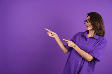 Young woman in a shirt with short sleeves on a purple background, with makeup and stylish glasses, points a finger to the left with a smile