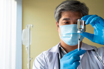 An Asian male doctor wears a medical glove and face mask holding syringe taking covid 19...