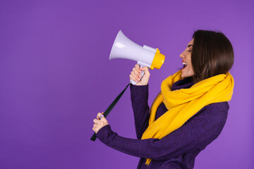 Young woman in a knitted dress and in a yellow scarf on a purple background cheerfully shouts into a megaphone, cheerful, announces a sale